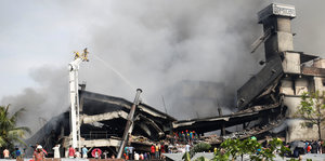 Ein Fabrikgebäude ist halb zerstört, Rauch steigt auf, im Vordergrund stehen Menschen