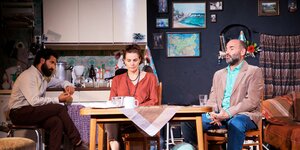 Szene aus Milo Raus „Empire“, drei Personen sitzen auf der Bühne in einer Küche
