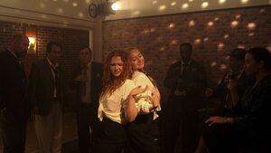 Zwei Frauen in weißen Hemden tanzen Rücken an Rücken in einem Club, um sie herum stehen Menschen und sehen zu
