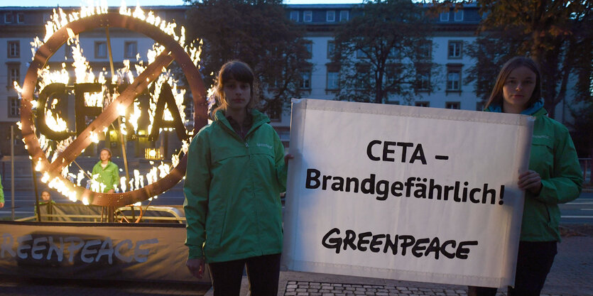 Ein brennendes Ceta-Symbol. Davor stehen zwei Frauen mit einem Plakat auf dem steht: Ceta – brandgefährlich. Darunter prangt der Greenpeace-Schriftzug