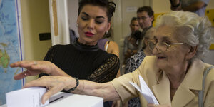 Eine ältere Frau steckt einen Zettel in eine Wahlurne, eine jüngere hilft ihr dabei