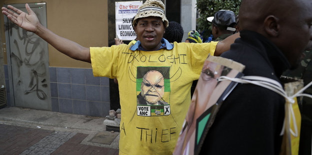 ein Mann in einem gelben T-Shirt breitet die Arme aus, auf dem T-Shirt ist ein Bild Zumas mit aufgemalten Teufelshörnern