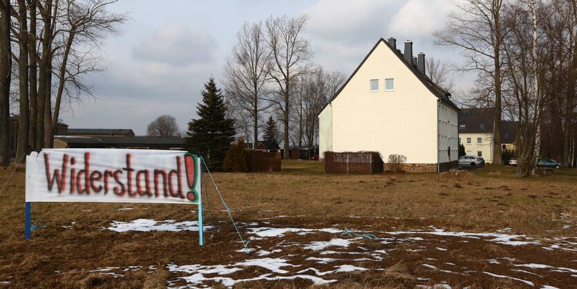 Ein Banner auf einer Wiese. Auf ihm steht „Widerstand“. Dahinter befindet sich ein Haus.