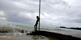 Ein Mensch lehnt an einem Pfahl, eine Welle Meerwasser schwammt daneben über den Strand