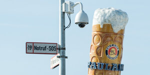 An einer Laterne hängt ein „Notruf“-Schild, daneben ragt ein künstlicher Bierkrug in die Höhe