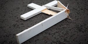 Ein zerbrochenes weißes Holzkreuz liegt auf dunklem Asphaltboden