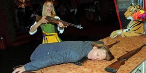 Dirndl-Trägerin hält Schießgewehr in den Händen. Vor ihr liegt ein Mann schlafend mit seinem Oberkörper auf einem Tresen