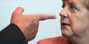 Eine Hand zeigt mit dem Zeigefinger auf Angela Merkels Nase