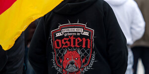 Auf einem schwarzem Pullover steht „Natürliche Härte geboren im Osten“, daneben hängt eine Deutschlandfahne
