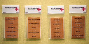 Vier Packungen mit der Aufschrift „Heldenhilfe“ hängen an einer gelben Wand. Sie enthalten Pflaster, auf denen Heldennamen stehen