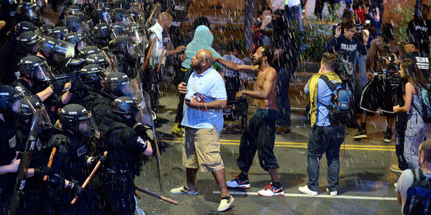 Links stehen viele Polizisten in voller Schutzkleidung, rechts stehen ihnen einzelne Menschen gegenüber, von oben kommen Wasserspritzer