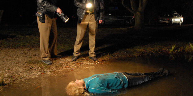 Es ist Nacht, in einer Pfütze liegt ein toter Mensch, der von zwei Polizisten mit Taschenlampen angeleuchtet wird