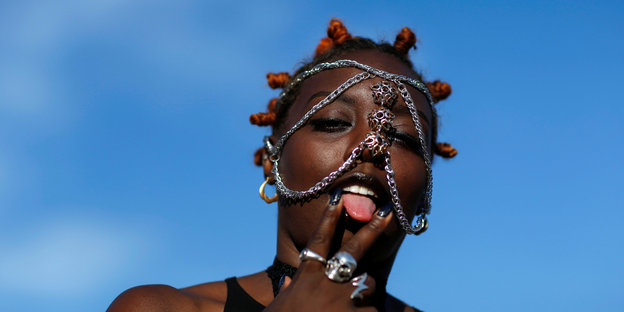 Eine Frau, deren Gesicht mit Piercings und Ketten geschmückt ist, streckt ihre Zunge zwischen zwei Fingern heraus