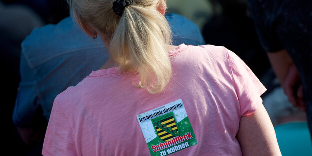 Frau von hinten, auf deren rosa T-Shirt steht: "Ich bin stolz darauf, im Schandfleck zu leben"