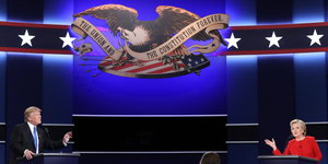 Donald Trump und Hillary Clinton stehen an Stehpulten, zwischen ihnen ein Symbol der USA: ein Adler mit einer Banderole, auf der „The Union and the Constitution forever“ steht