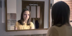 Eine blasse junge Frau mit blutunterlaufenen Augen schaut in einen Spiegel