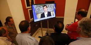Auf einem Fernsehbildschirm ist Alexis Tsipras zu sehen, davor stehen eine Reihe Mensche im Halbkreis