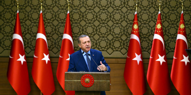 Ein Mann in Anzug steht hinter einem Podium und spricht in ein Mikrofon, hinter ihm sind türkische Nationalflaggen aufgestellt