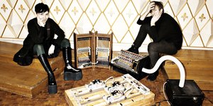 Die schwarzgekleideten Marion Wörle und Maciej Śledziecki sitzen, zwischen und vor ihnen stehen mehrere aus Holz gebaute, teils verkabelte Maschinen