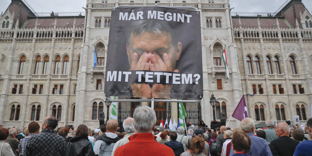In einer Menschenmenge trägt eine Person ein Plakat auf dem Viktor Orban das Gesicht in den Händen begräbt