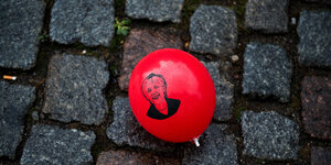 Ein roter Luftballon mit dem Porträt Merkels liegt auf grauem Kopfsteinpflaster