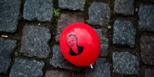 Ein roter Luftballon mit dem Porträt Merkels liegt auf grauem Kopfsteinpflaster