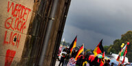 Ein rotes Grafitti „Werdet glücklich“, schräg dahinter Menschen mit Deutschlandfahnen