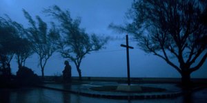 Vor einem dunklen Himmel steht zwischen zerzausten Bäume und einem dunklen Kreuz ein Mensch
