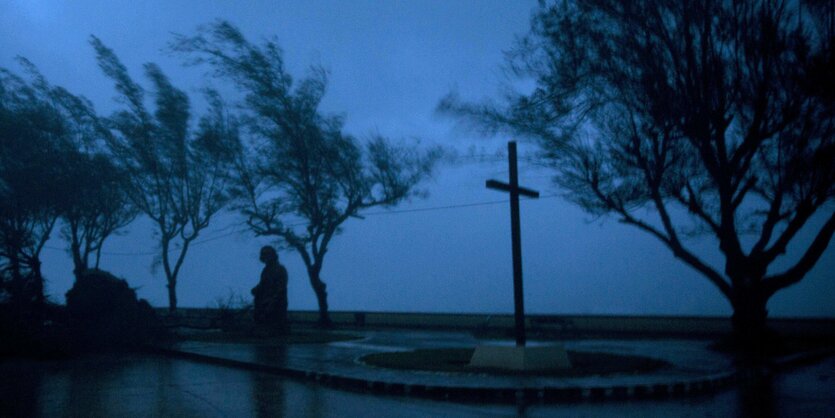 Vor einem dunklen Himmel steht zwischen zerzausten Bäume und einem dunklen Kreuz ein Mensch