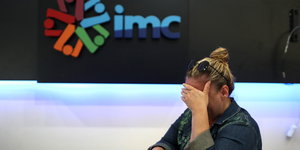 Eine Frau hält sich mit gesenktem Kopf die Hand vor die Augen, hinter ihr das IMC-Logo