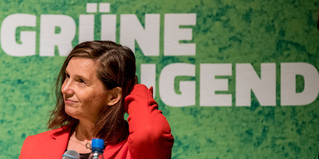 Die grüne Fraktionsvorsitzende Katrin Göring-Eckardt steht in einem roten Blazer vor einer Grünen Wand mit der Aufschrift "Grüne Jugend"