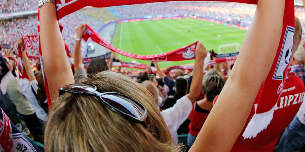 Eine Frau hält einen Fan-Schal vom Fußballklub RB Leipzig hoch. Zwischen ihren Armen sieht man das Stadion