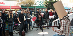 Ein Mann mit lustigem Papphut spielt vor einer Menge Menschen Schlagzeug auf der Straße