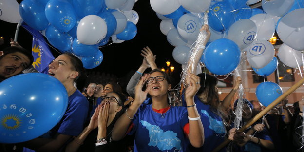 Mehrere junge Menschen in blauen T-Shirts applaudieren und halten mit Hellium gefüllte, blaue und weiße Luftballons