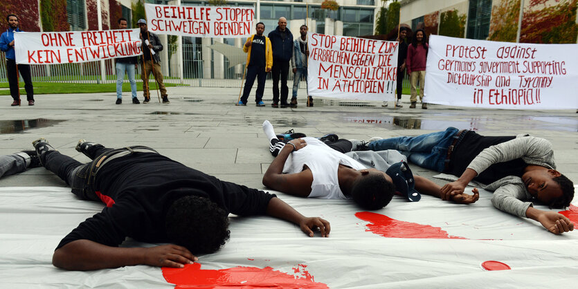 Auf einer weißen Plane liegen Menschen, rote Farbe ist neben ihren Köpfen verteilt, dahinter stehen Demonstraten_innen mit Transparenten