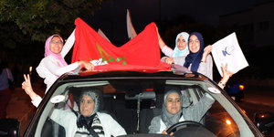 Fünf Frauen, die Kopfücher tragen, sitzen in einem Auto am Steuer und aus den Fenster gelehnt, zeigen das Victory-Zeichen und halten Fahnen
