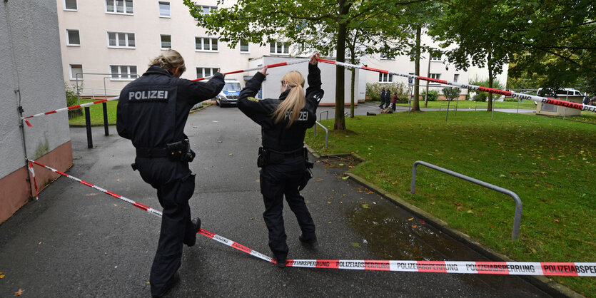 Zwei Polizisten gehen unter einem Absperrband hindurch zu einem Wohnhaus