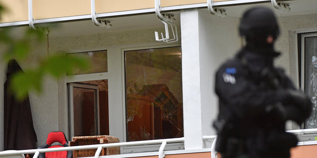 Ein komplett schwarz gekleideter Polizist, hinter ihm ein Balkon, die Balkontür ist teils zersplittert