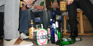 Mehrere Menschen sitzen auf Stühlen in einer Art Halbkreis, in der Mitte sind auf dem Boden viele Alkohol-Flaschen