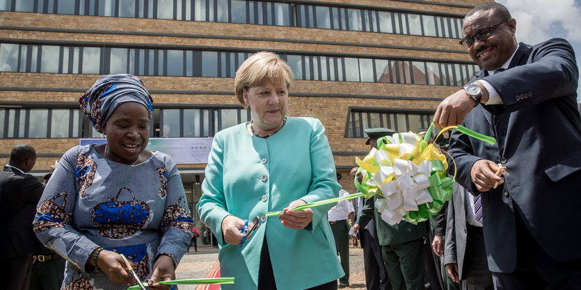 Von links nach rechts: Eine schwarze Frau mit Kopfbedeckung, eine weiße Frau in hellblauem Anzug und ein schwarzer Mann in Anzug schneiden ein feierliches Band durch