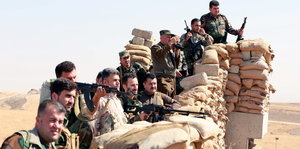 Inmitten einer Wüstenlandschaft stehen Soldaten mit Waffen vor einer Mauer aus Sandsäcken