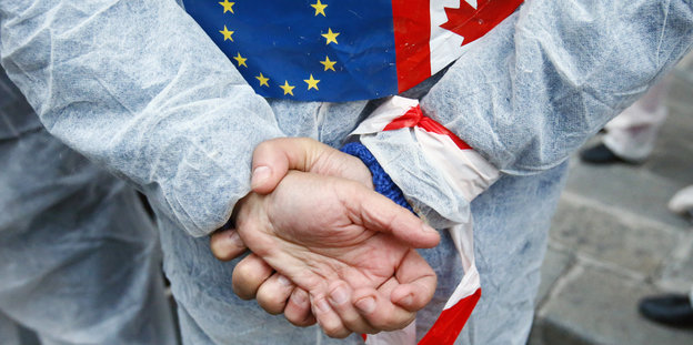 Eine Person hält seine Hände hinterm Rücken zusammen. An seinem Rücken sind auch die Flaggen von EU und Kanada