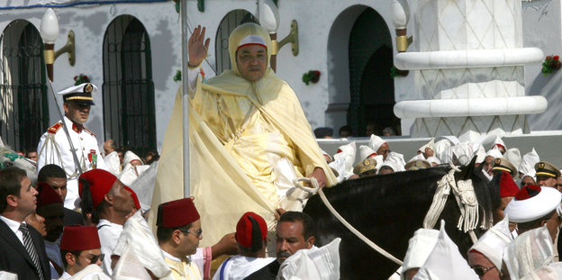 Mohammed VI. reitet auf einem Pferd unter einem roten Sonnenschirm durch eine Menschenmenge