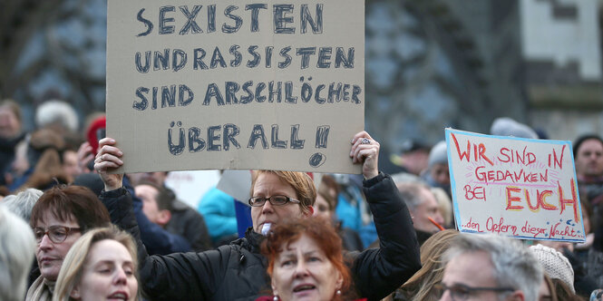 Ein Frau hält ein Schild hoch „Sexisten und Rassisten sind Arschlöcher. Überall!", um sie herum stehen Menschen