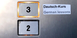 Zwei Aufzugknöpfe mit den Nummern 2 und 3. 3 ist erleuchtet, daneben ein Schild „Deutsch-Kurs - German lessons“