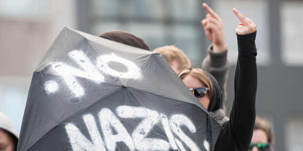 Personen halten einen Regenschirm mit der Aufschrift "No Nazis" hoch und heben ihre Hände zu obszönen Gesten