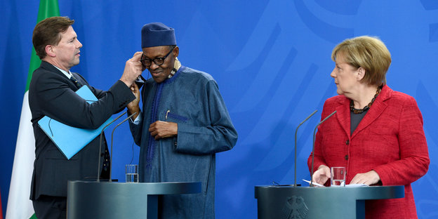 Steffen Seibert setzt Nigerias Präsident Buhari Kopfhörer auf während Angela Merkel daneben steht