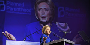 Eine Frau mit kurzen blonden Haaren steht an einem Redepult und spricht ins Mikrofon, im Hintergrund wird sie live groß auf eine Leinwand projiziert