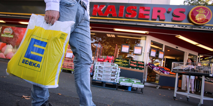 Vor einem Supermarkt mit der Aufschrift Kaiser's geht jemand mit einer gelben Edeka-Tüte vorbei