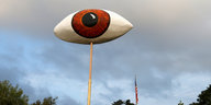 Ein Pappmaché-Auge ragt in den Himmel, weit dahinter eine USA-Fahne